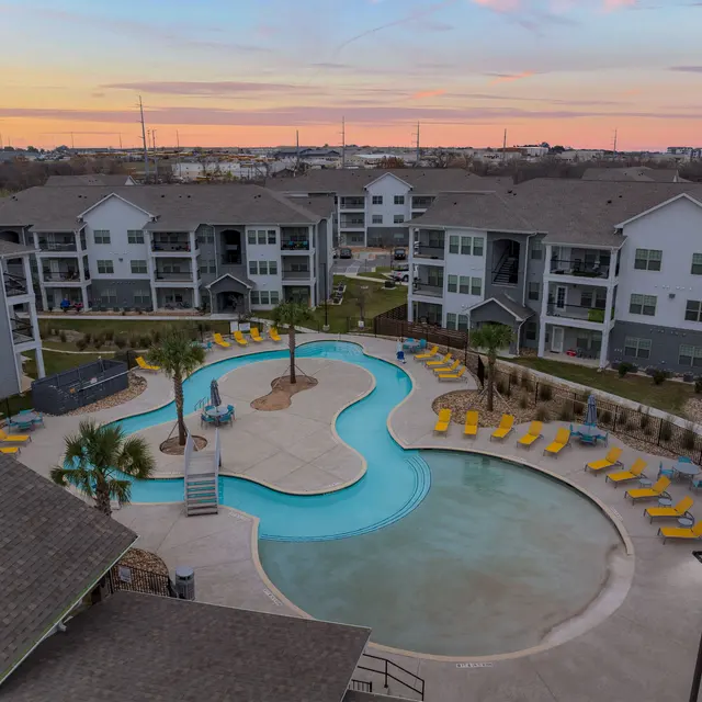 Aerial view of a modern apartment complex with a uniquely shaped swimming pool surrounded by lounge chairs. The sky is vibrant, showing hues of orange and purple at sunset.