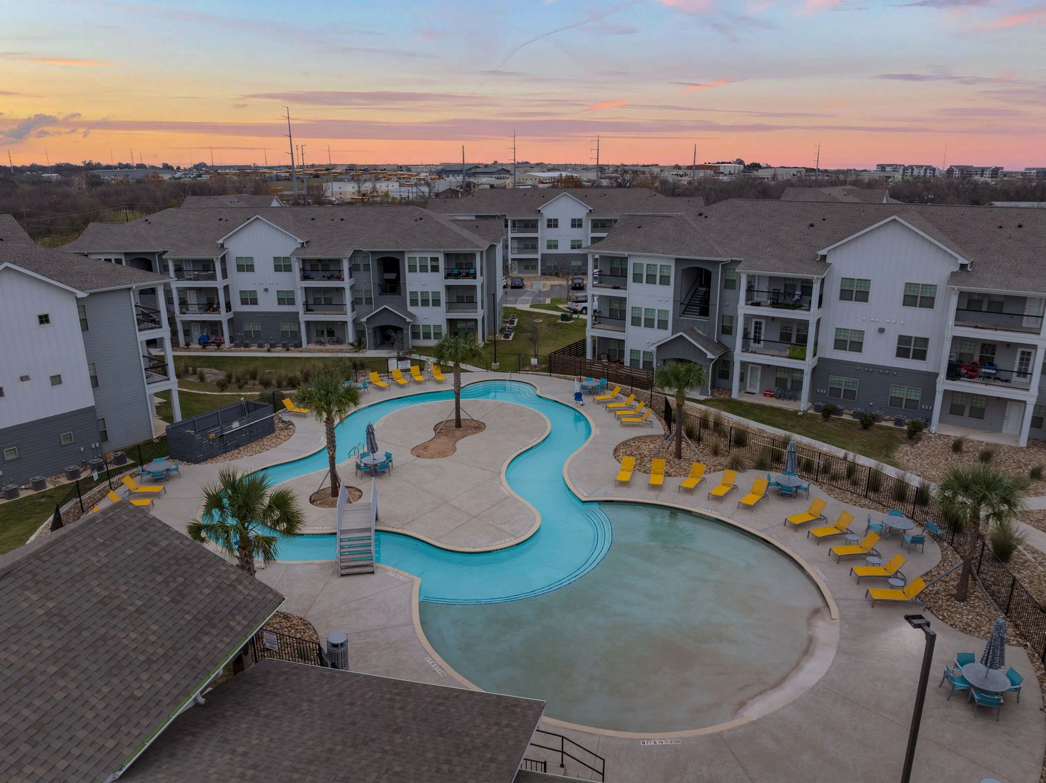 Aerial view of a modern apartment complex with a uniquely shaped swimming pool surrounded by lounge chairs. The sky is vibrant, showing hues of orange and purple at sunset.