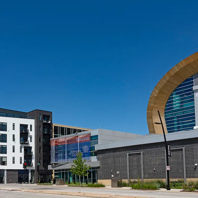 A wide shot of a street featuring a modern urban building on the left side and the exterior of a large arena, Fiserv Forum, on the right. The sky is clear and blue.
