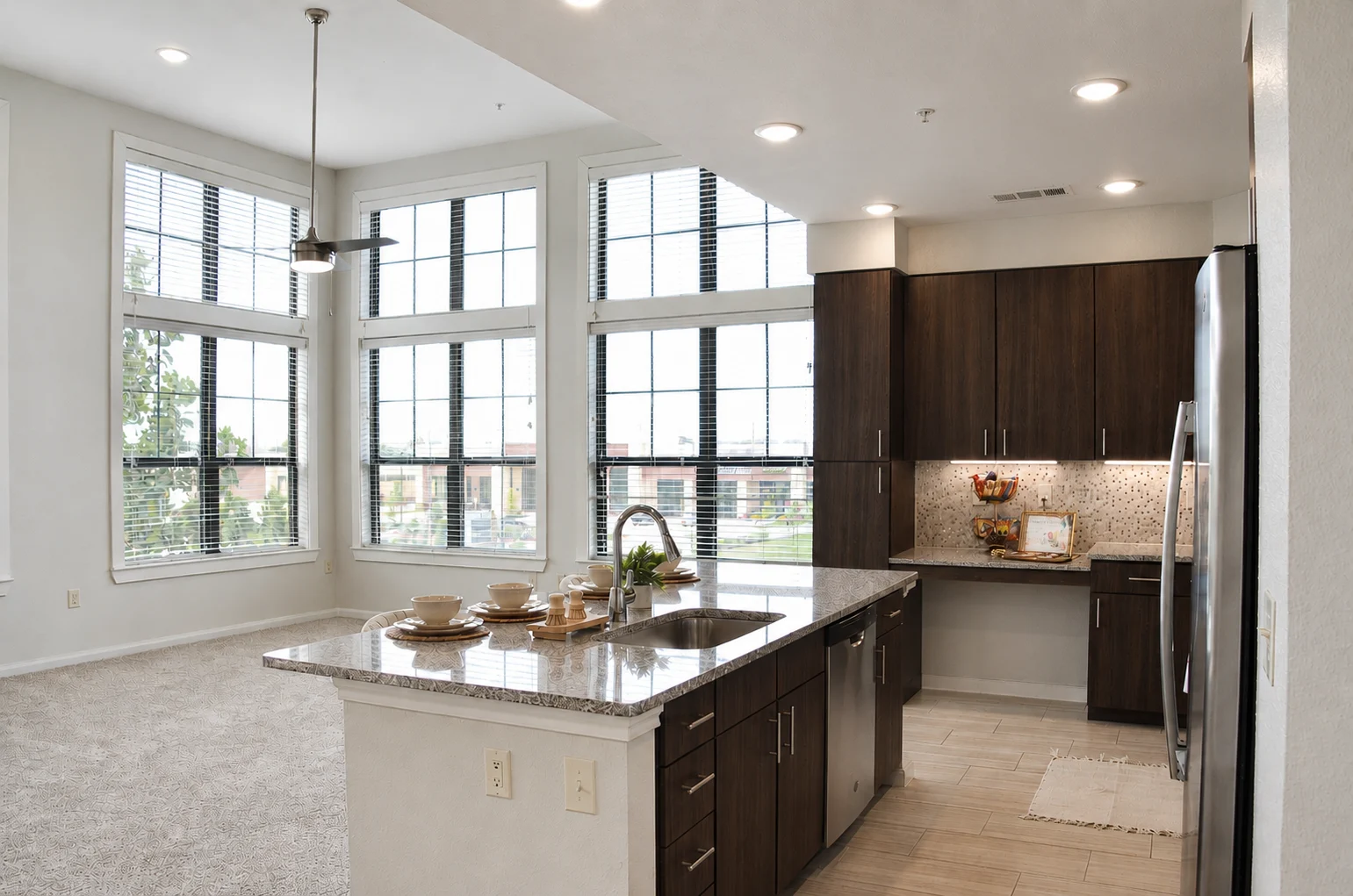 Modern Kitchen and Living Area A modern kitchen with dark cabinets, granite countertop, and a large window overlooking a living space with carpet and bright lighting.