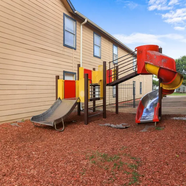 A small playground area featuring two slides and a climbing structure surrounded by red mulch, located between two buildings.