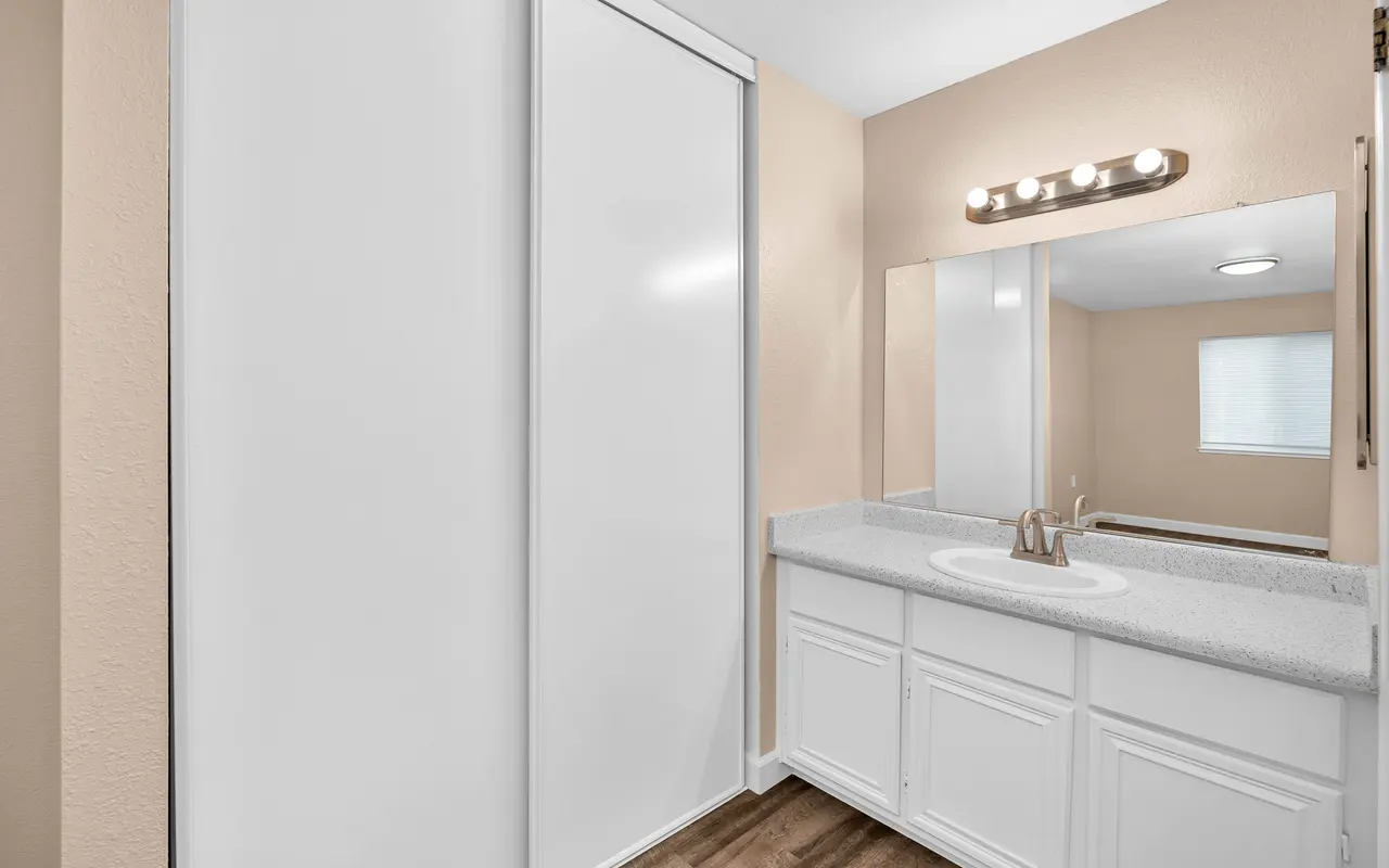 A modern bathroom featuring a white sink with a faucet, a large mirror above it, and light brown wood-like flooring. There are sliding doors to the left, and a light fixture with multiple bulbs above the mirror. A window allows natural light into the space.
