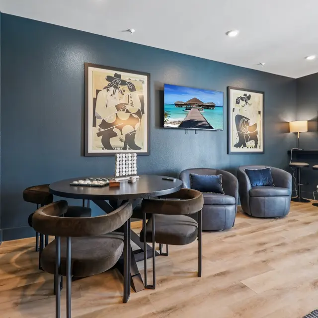 Modern Living Space with Artistic Touch A modern living area featuring a round table with four black chairs, two plush blue armchairs, and framed artwork on the wall. There is a long mirror and bar stools along another wall, with a stylish lamp providing light.