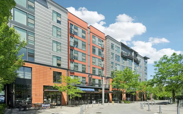 A modern apartment building with a mix of orange and gray panels, featuring ground-level retail spaces and surrounded by trees under a clear sky.