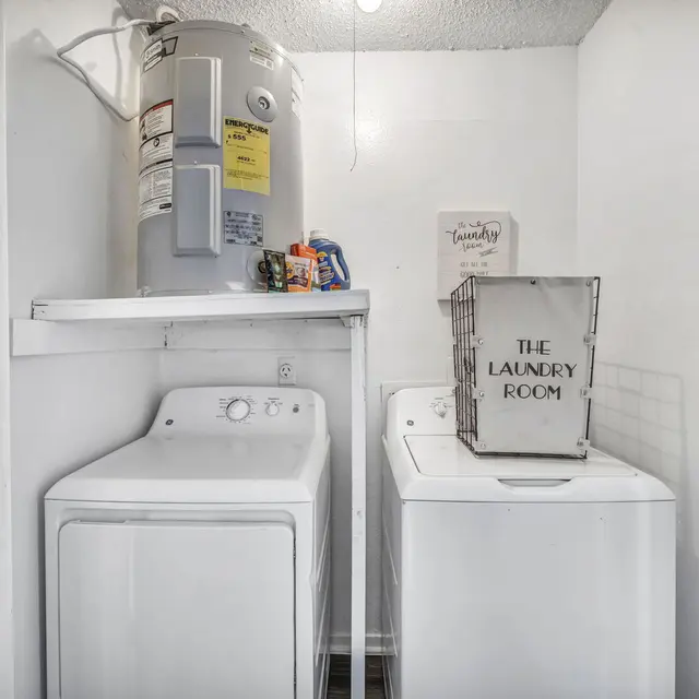 A laundry room with a washer and dryer, shelves with cleaning supplies, and a basket labeled "THE LAUNDRY ROOM."