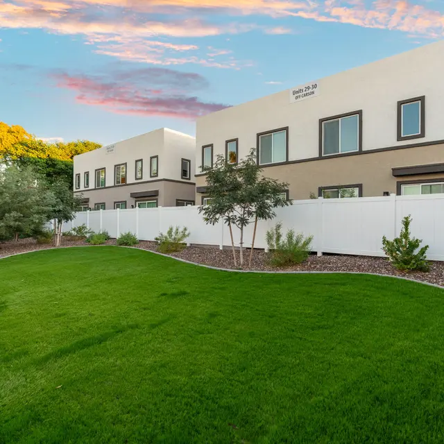 Modern Landscape with Green Lawn and Contemporary Buildings A green lawn with a curved design, surrounded by a clean white fence and modern buildings in the background under a colorful sky at dusk.