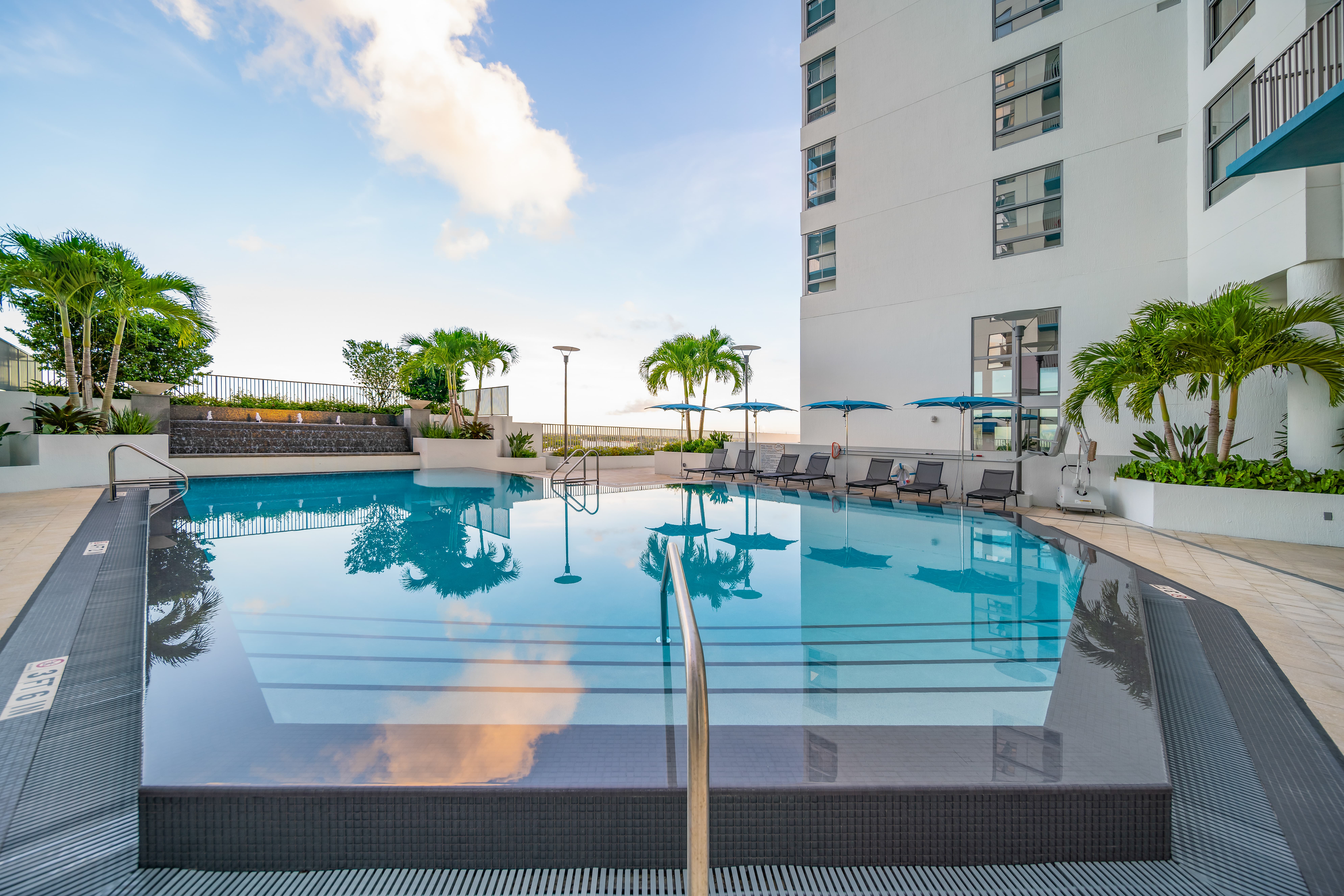 A modern swimming pool area surrounded by palm trees and lounge chairs, with a clear blue sky in the background.