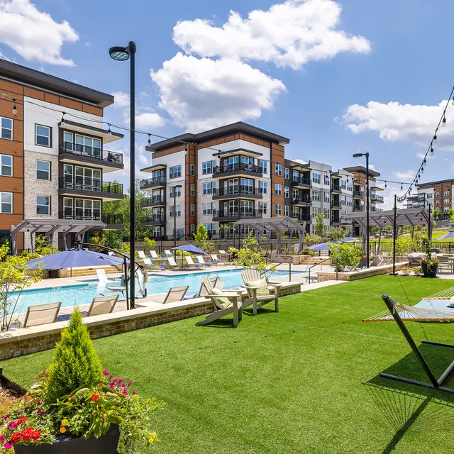 A picturesque view of a modern apartment complex featuring a swimming pool surrounded by lounge chairs and green landscaping.