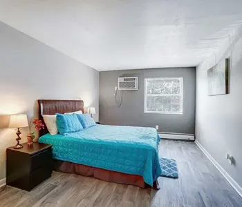 A modern bedroom with a queen-sized bed dressed in a blue quilt. There is a nightstand with a lamp beside the bed and a window with natural light coming in. The walls are painted a light gray, and the floor is made of wood.