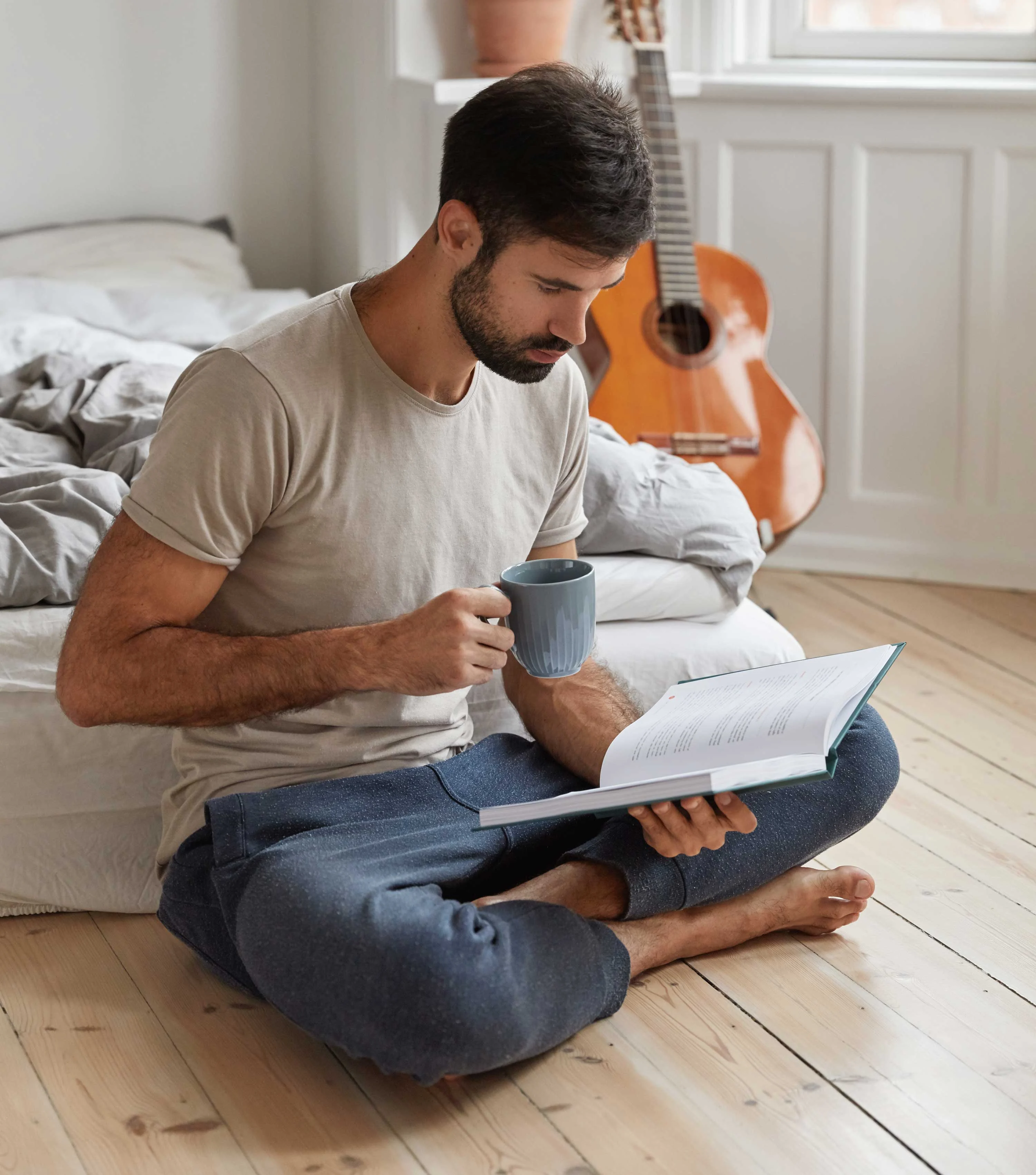 Man Reading a Book A man sitting cross-legged on a wooden floor, holding a cup of coffee and reading a book. He is wearing a beige t-shirt and gray sweatpants, with a bed in the background and an acoustic guitar leaning against the wall.