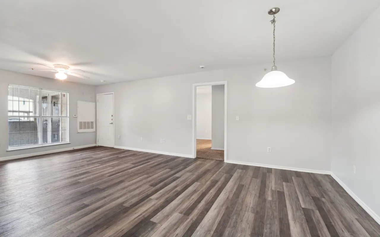 A spacious and bright living room featuring wooden flooring, a ceiling fan, and a modern light fixture. The walls are painted in a light color, and there are doorways leading to other rooms. Large windows provide natural light.