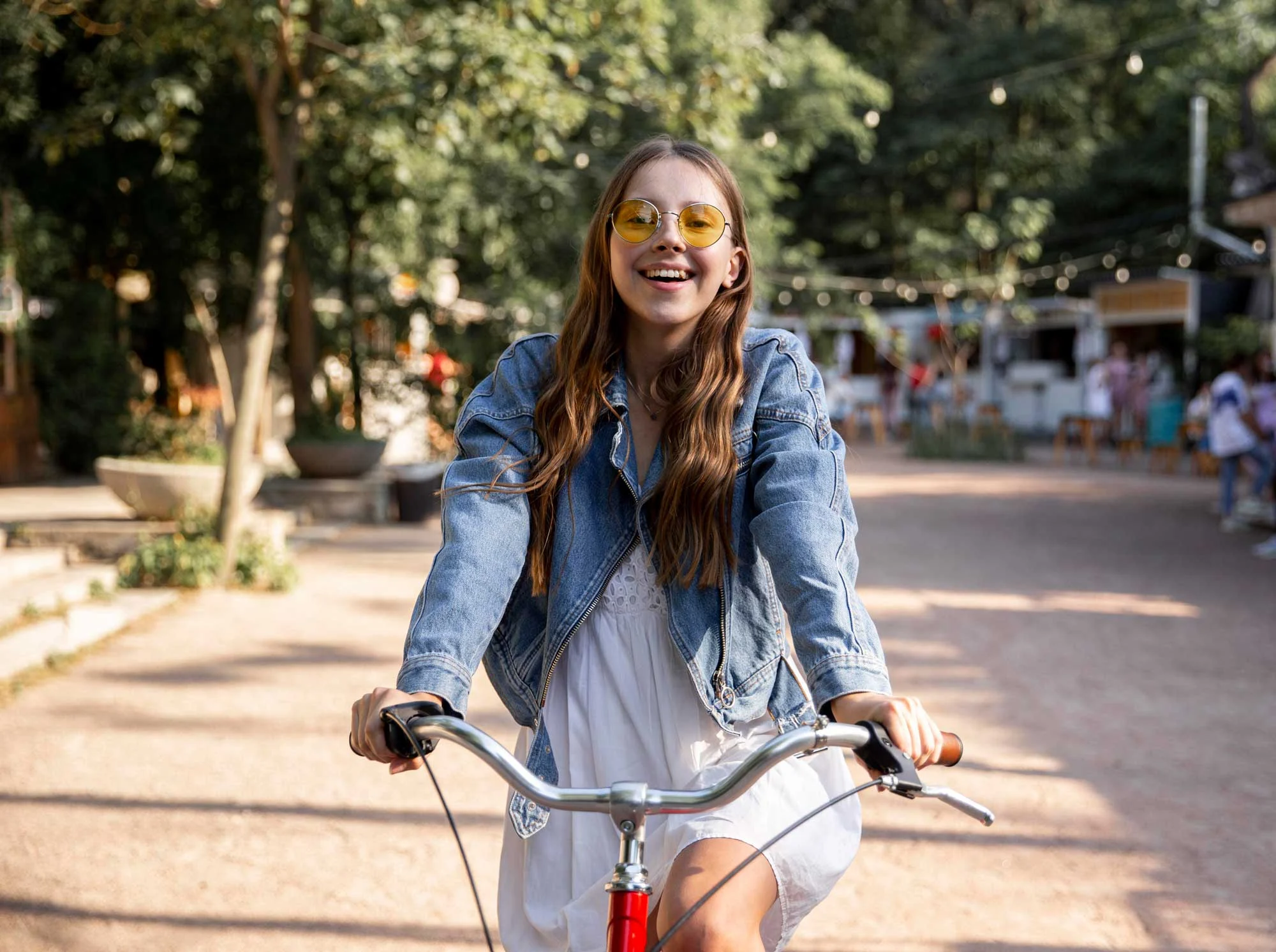 Joyful Bike Ride in the Park A young woman with long brown hair wearing a denim jacket and yellow sunglasses, smiling while riding a bicycle in a park-like setting. She is dressed in a white dress and the background features greenery and soft lighting.