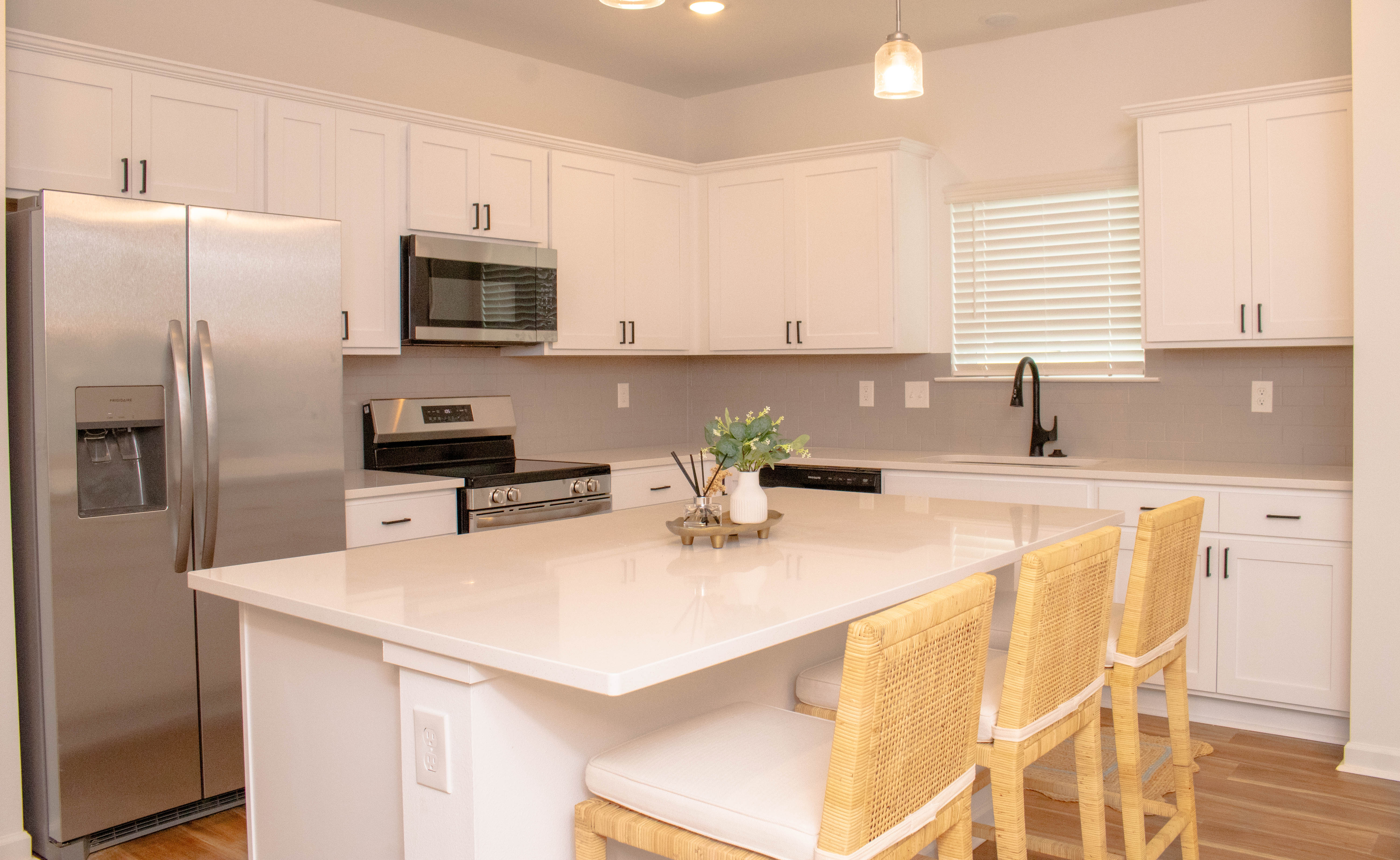 A modern kitchen featuring white cabinetry, stainless steel appliances, and a large island with seating. The kitchen is bright with natural light coming from a window, complemented by minimalist decor.