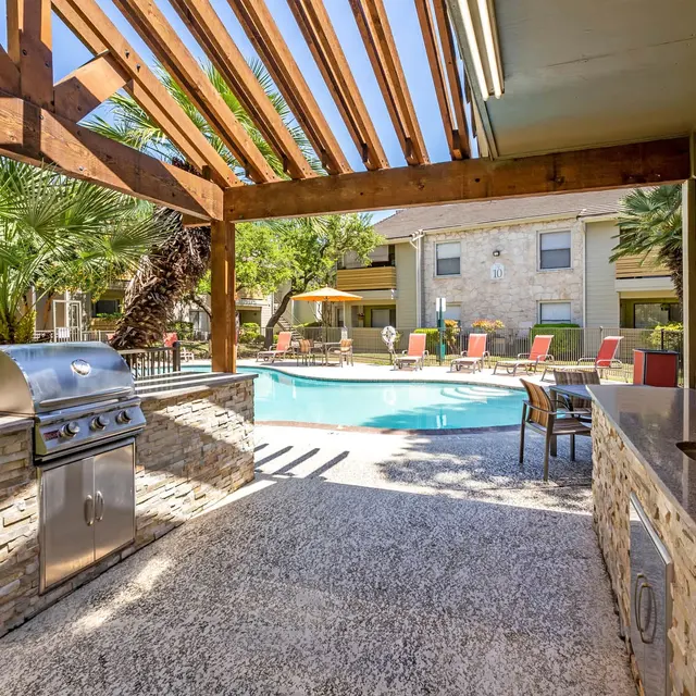 Outdoor pool area with a barbecue grill and seating under a wooden pergola, surrounded by palm trees.