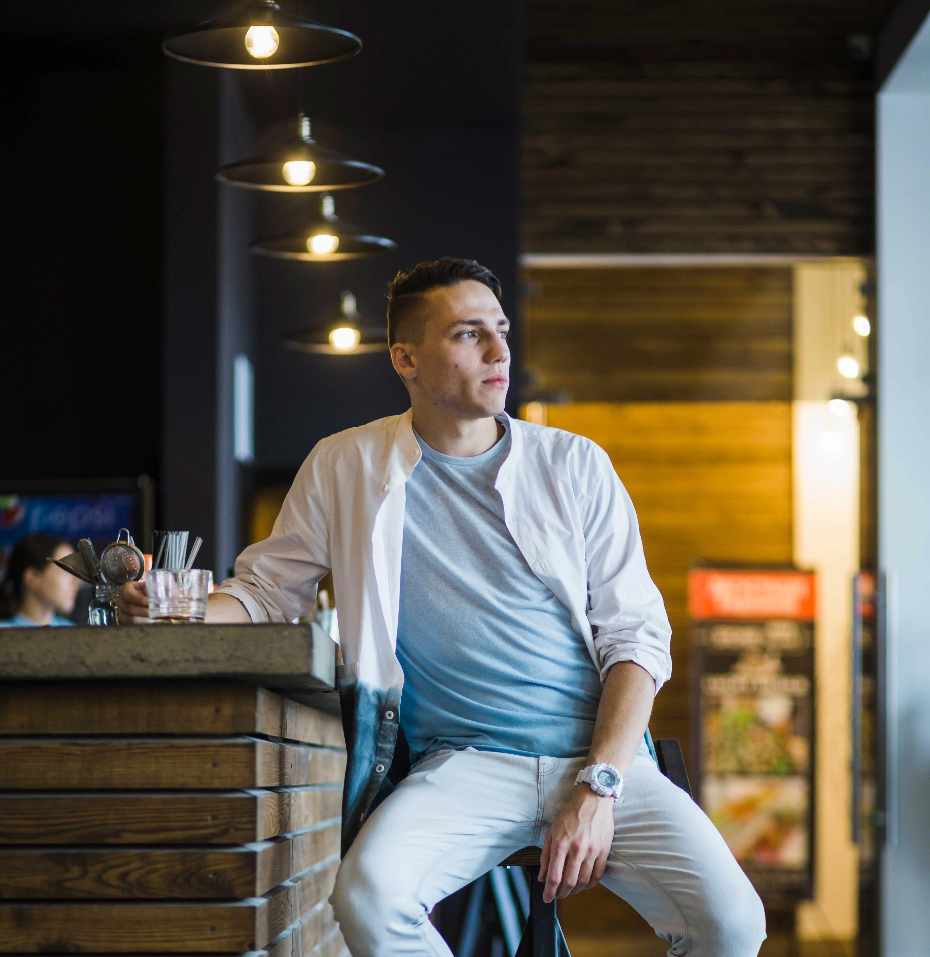 Moment in a Modern Café A young man sitting at a bar counter in a café, wearing a light shirt and gray t-shirt, with a drink in hand. The atmosphere is modern and stylish, with warm lighting and wooden accents.