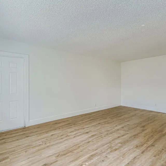 A spacious empty living room with light wooden flooring and white walls. A door is visible on the left side, and a stainless steel refrigerator is partially visible in an adjacent room.