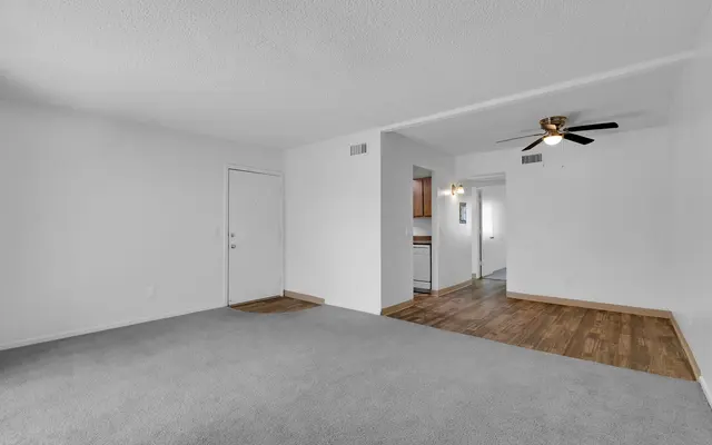 A spacious living room featuring light gray carpet and white walls. There is a doorway leading to another room and a view into a kitchen area with wooden floors in the background. A ceiling fan is visible in the right corner.