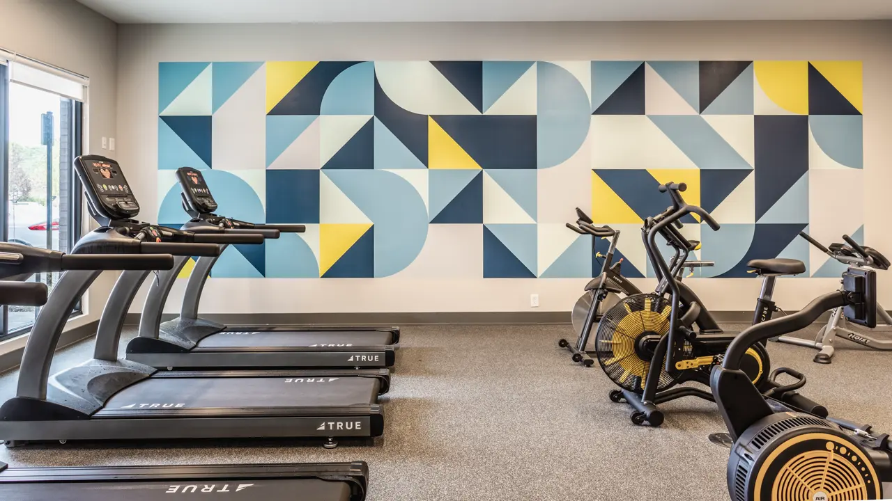 A modern gym interior featuring several treadmills and exercise bikes, with a colorful geometric mural on the wall.