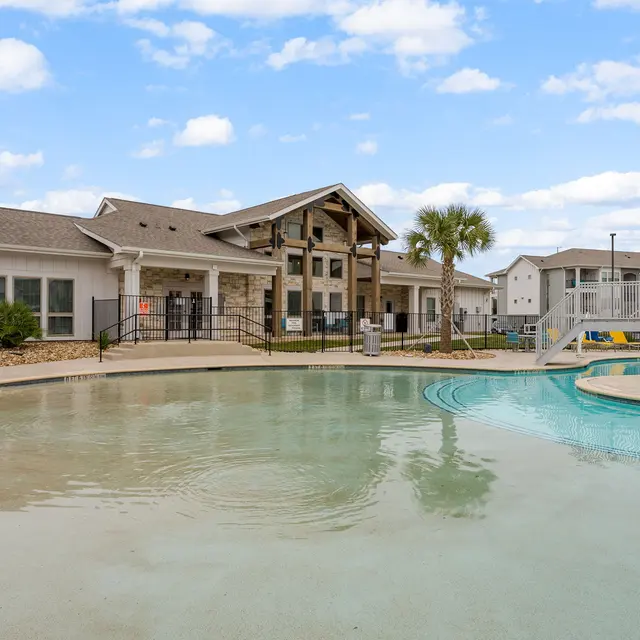 View of a residential apartment complex with a swimming pool, featuring a bridge and landscaped surroundings under a blue sky.