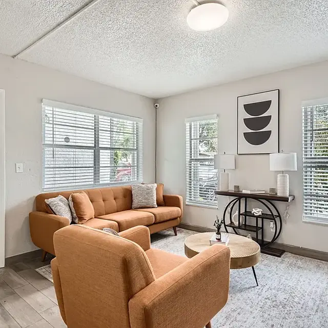 A cozy modern living room featuring an orange couch, a round coffee table, and large windows with blinds. The space is bright and airy with a neutral color palette and a simple decorative artwork on the wall.