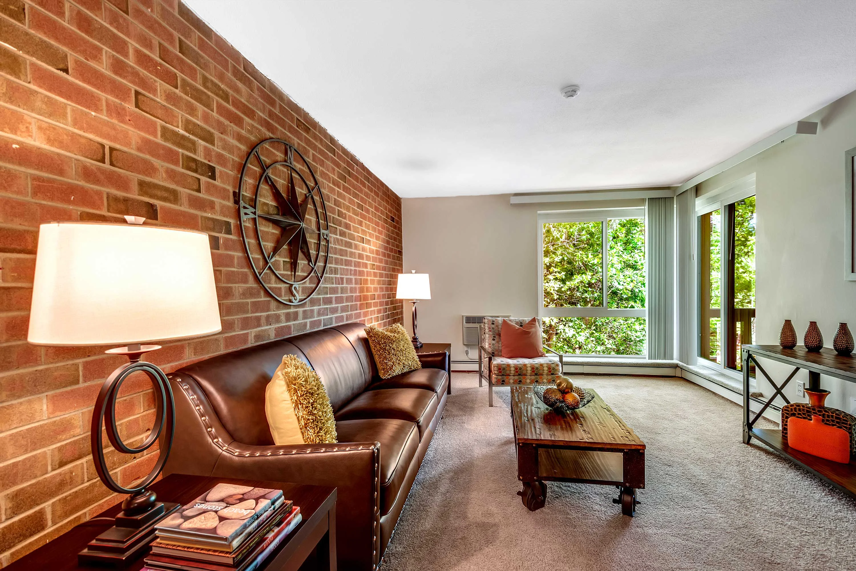 A cozy living room featuring a leather couch, a rustic coffee table, a large window with natural light, and a brick accent wall.