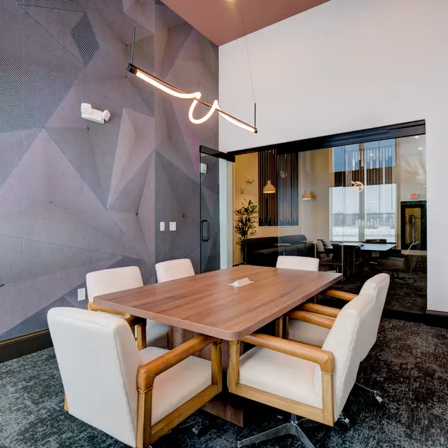 A modern meeting room featuring a wooden table surrounded by four white chairs. The walls are decorated with a geometric pattern, and there is a glass wall leading to another area. A pendant light hangs from the ceiling, illuminating the space.
