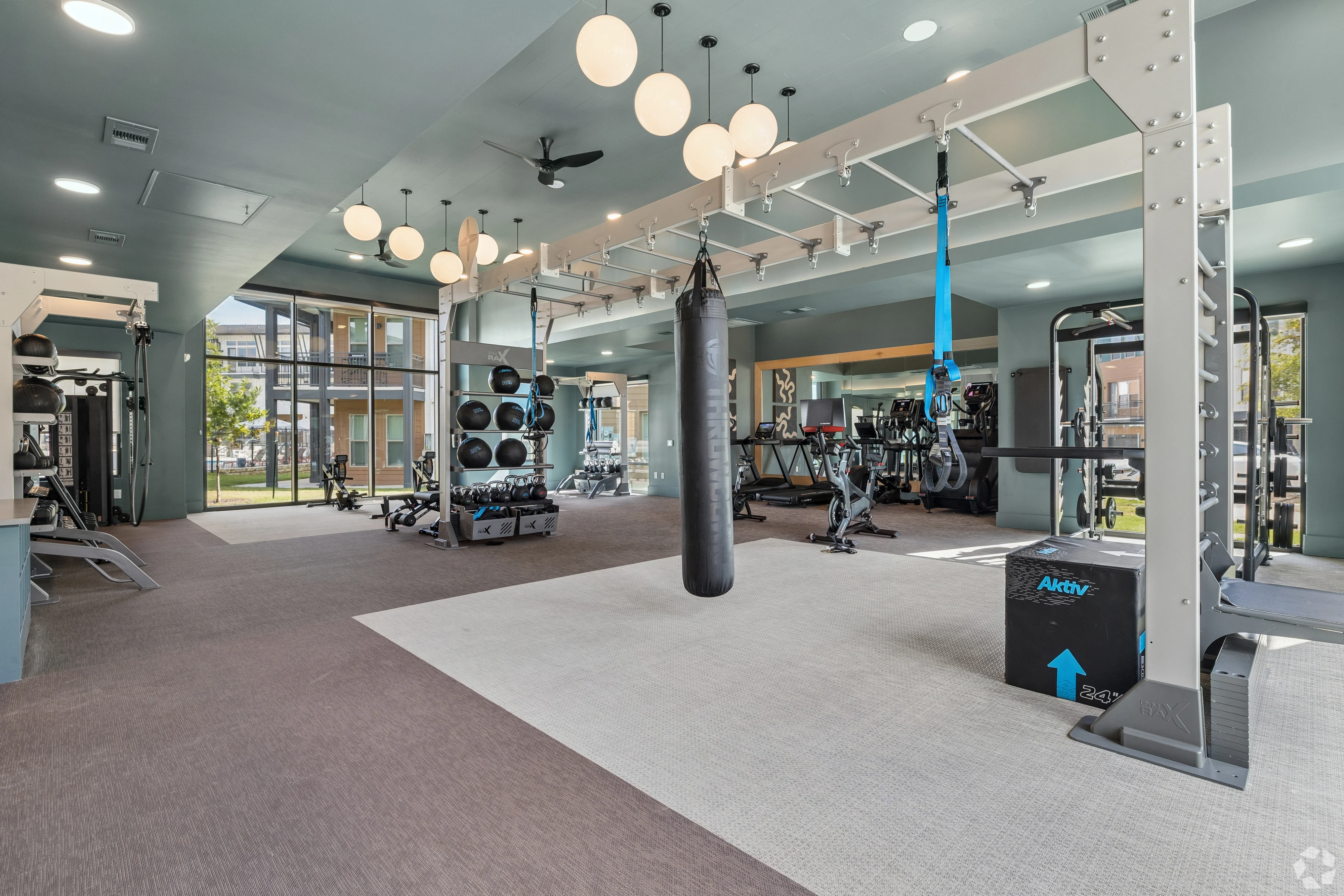 A spacious gym area with exercise equipment, including weights and a punching bag, large windows allowing natural light, and modern lighting fixtures.