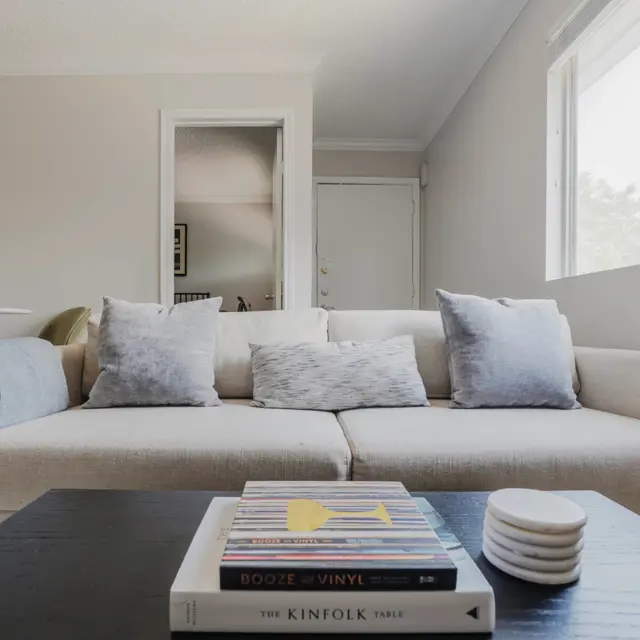 A cozy living room featuring a beige sofa with blue accent pillows, a dark wooden coffee table topped with books and cups, and a light-filled space with a neutral color palette.