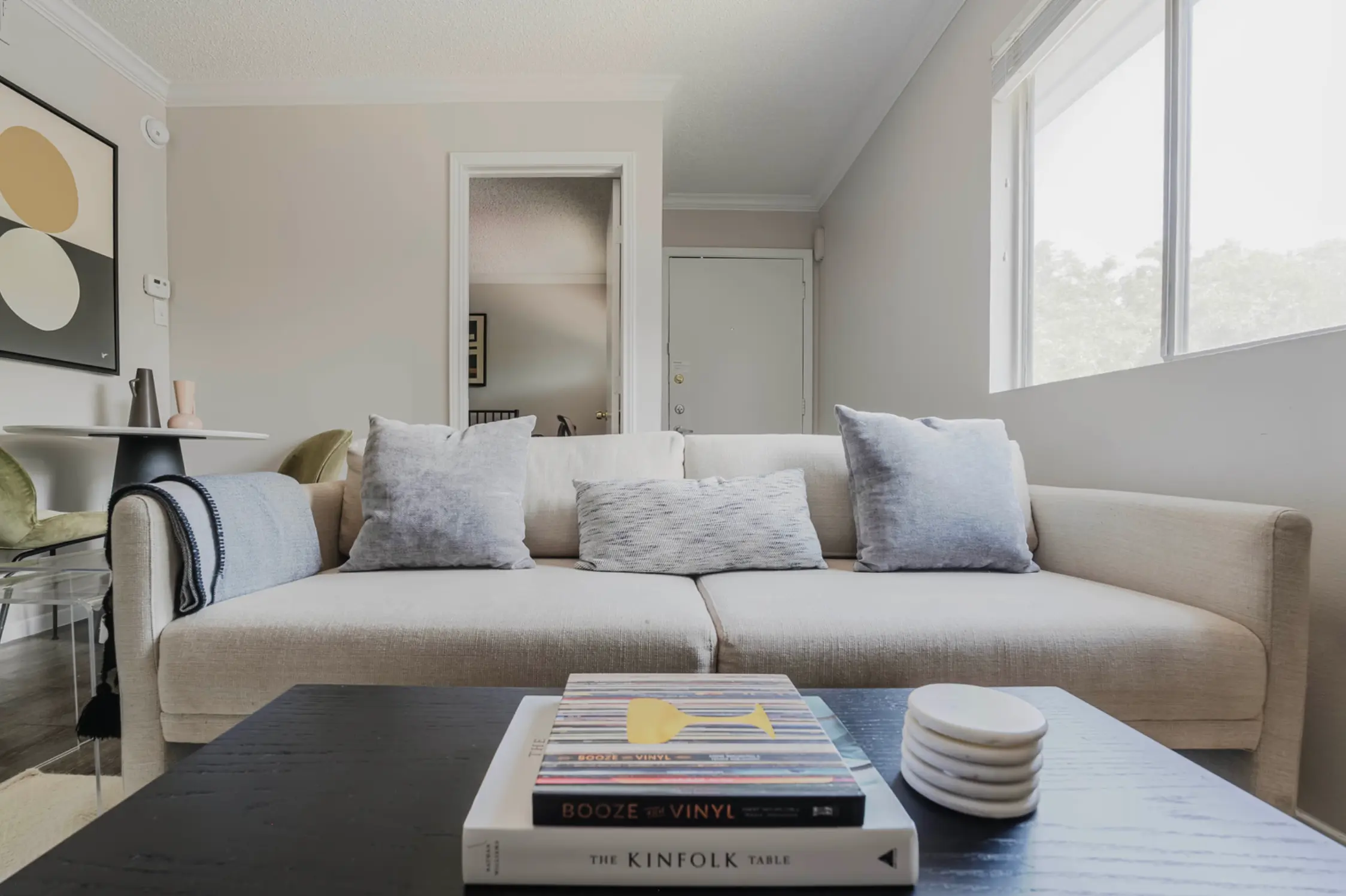 A cozy living room featuring a beige sofa with blue accent pillows, a dark wooden coffee table topped with books and cups, and a light-filled space with a neutral color palette.