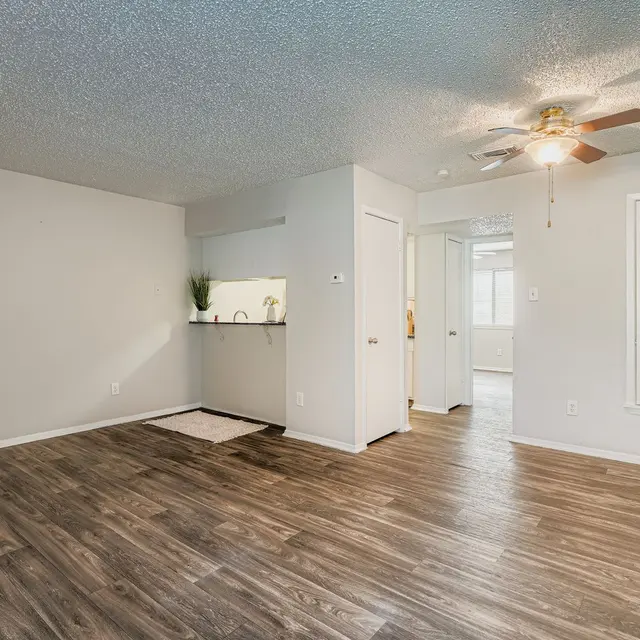 A spacious living room featuring wooden floors, a ceiling fan, and a doorway leading to another room. The walls are painted in a light color, and there is an open kitchen area visible in the background.