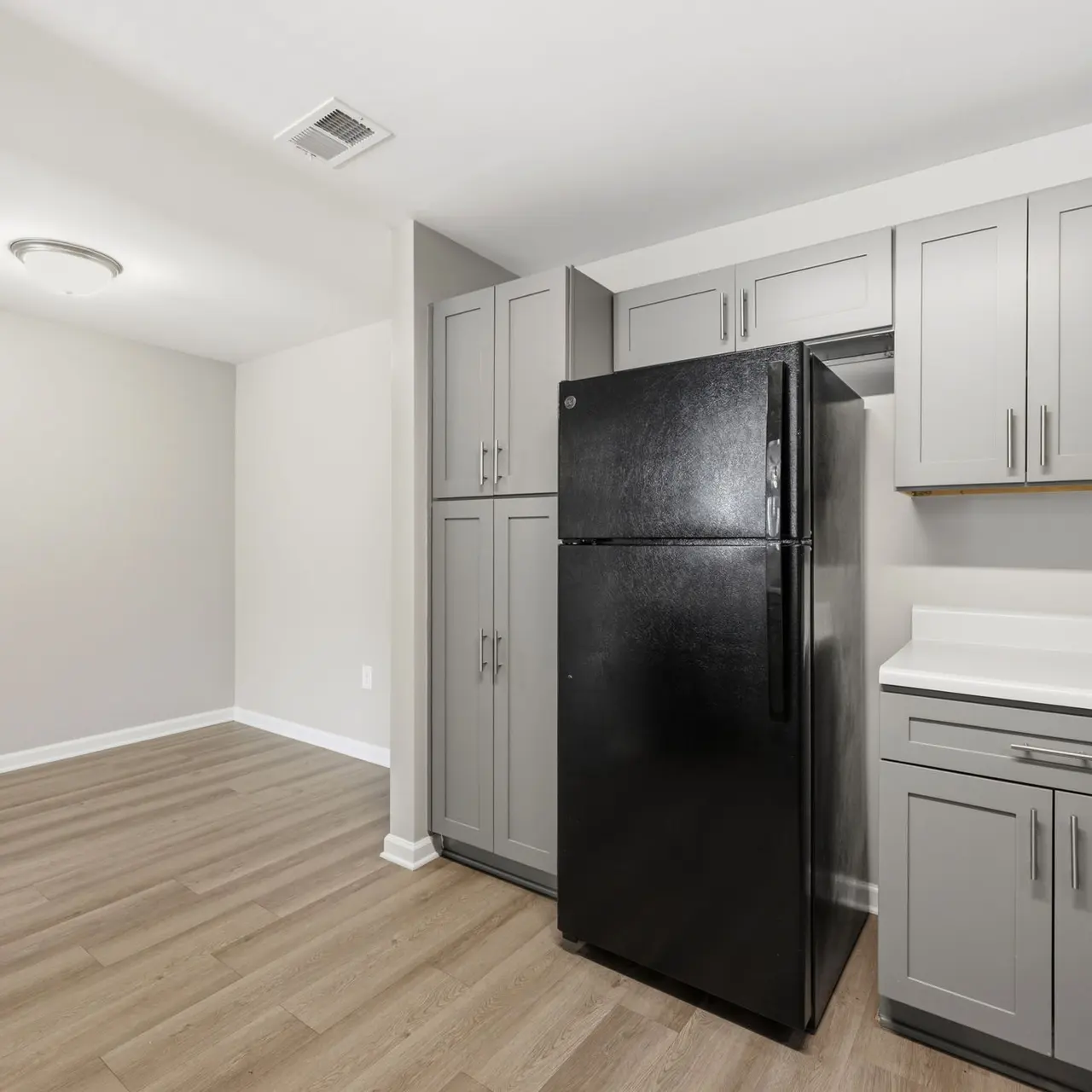 A modern kitchen featuring a black refrigerator, gray cabinets, and light hardwood flooring. The kitchen has a spacious layout with a counter and cabinetry.