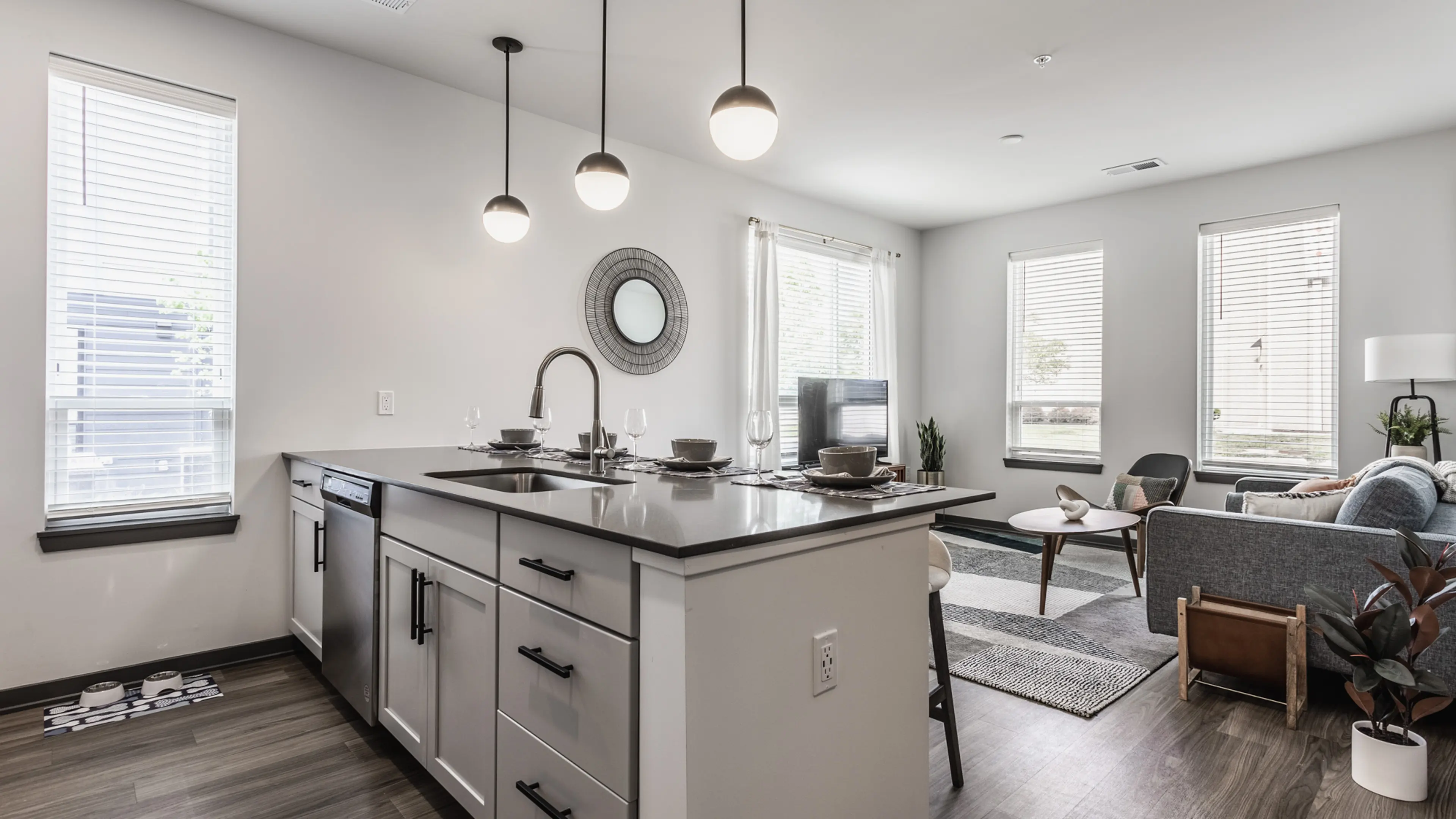 A modern open-concept kitchen and living area featuring a dark countertop, sleek cabinets, dining setup, stylish lights, and large windows with natural light.