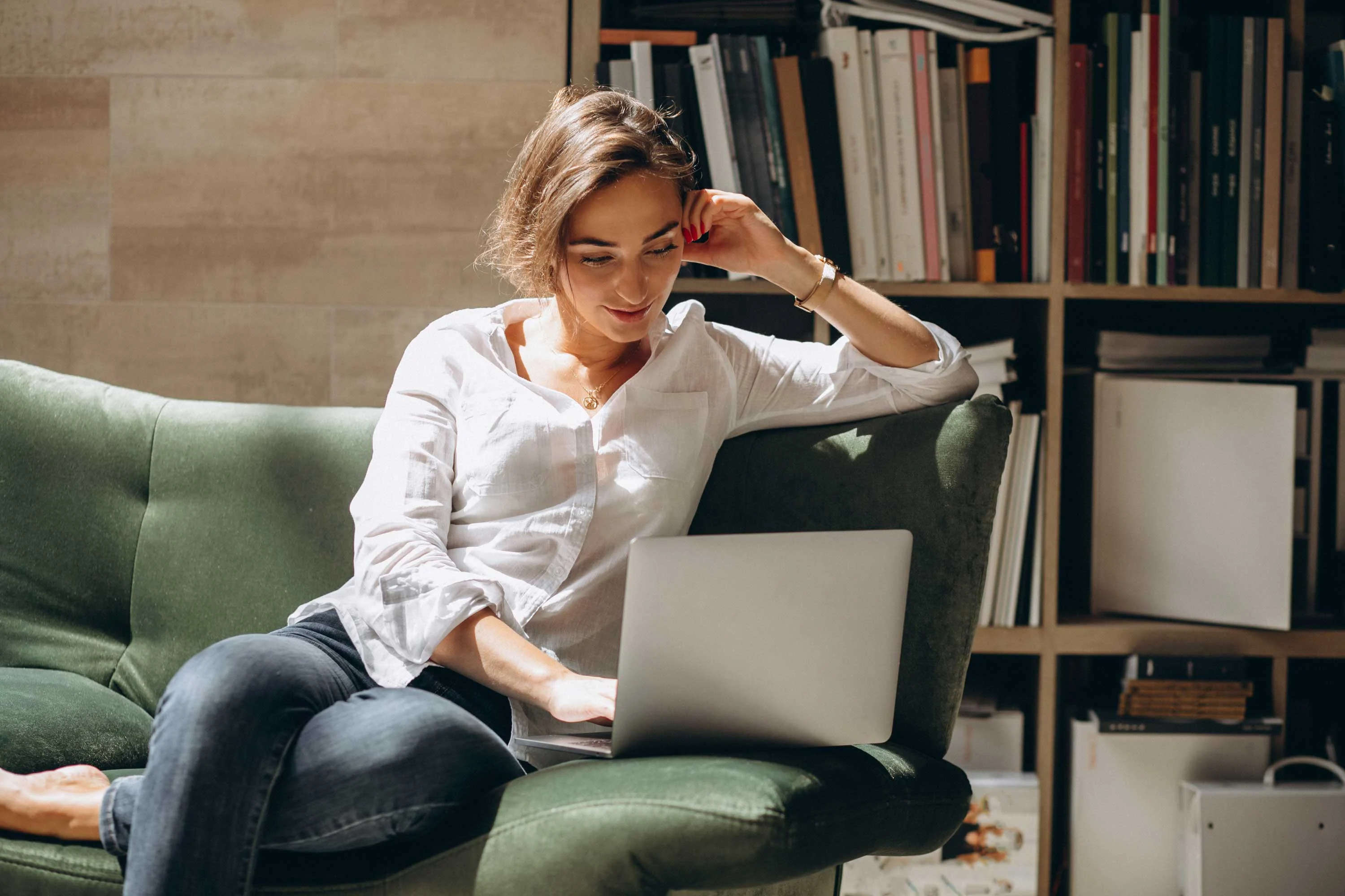 Home Workspace A woman sitting on a green couch, looking at a laptop, with a shelf of books in the background.