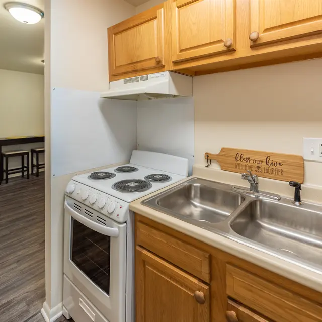 A small kitchen featuring wooden cabinetry, a white stove with four burners, a double sink, and a wooden sign that reads 'love grows here'.