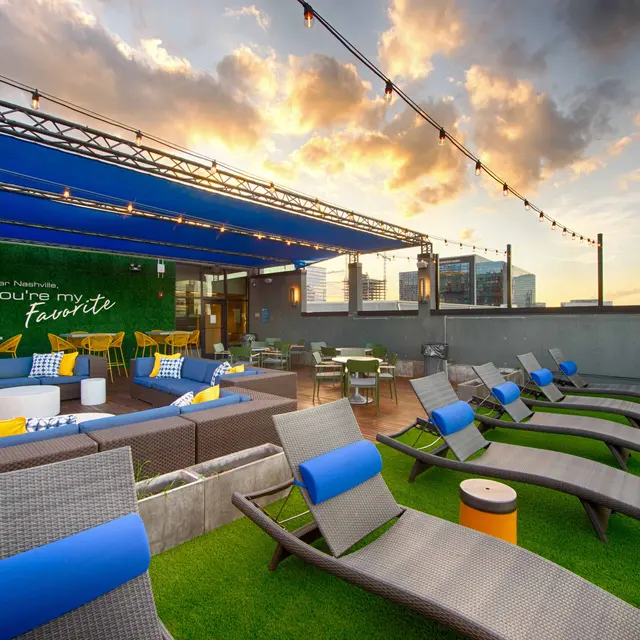A modern rooftop lounge featuring comfortable seating, vibrant cushions, and green grass. The area is adorned with decorative lighting and a colorful mural that reads 'You're my favorite.'
