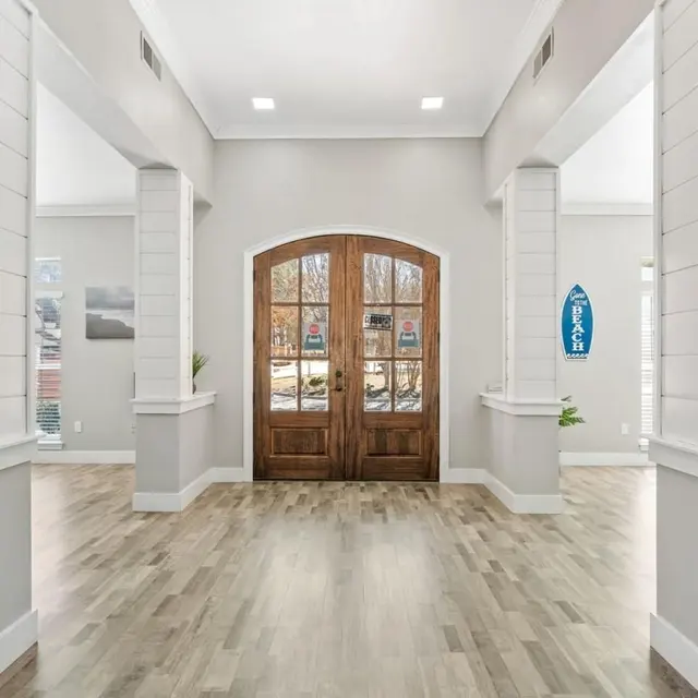 A spacious entryway featuring a wooden double door and large windows, with light-colored walls and wooden flooring.