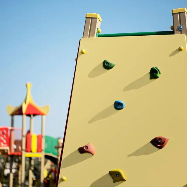 Colorful Playground Climbing Wall A bright yellow climbing wall with colorful holds in a playground setting, featuring a colorful play structure in the background under a clear blue sky.