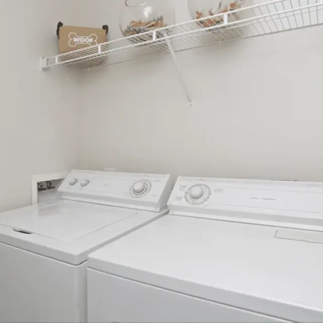 A laundry room featuring a washing machine and dryer side by side against a light-colored wall. Above them, there is a wire shelf holding decorative items.