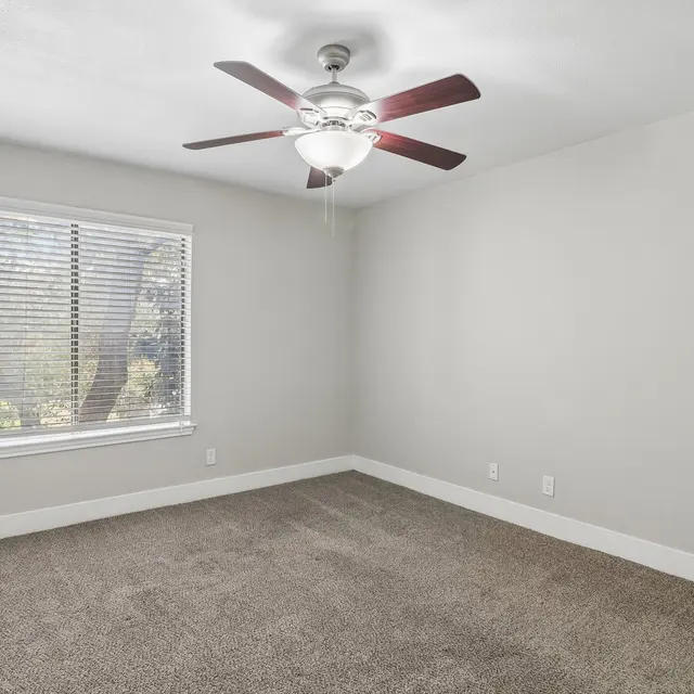 Vacant Bedroom with Ceiling Fan A vacant bedroom featuring a ceiling fan, large window with blinds, and neutral-colored walls and carpet.