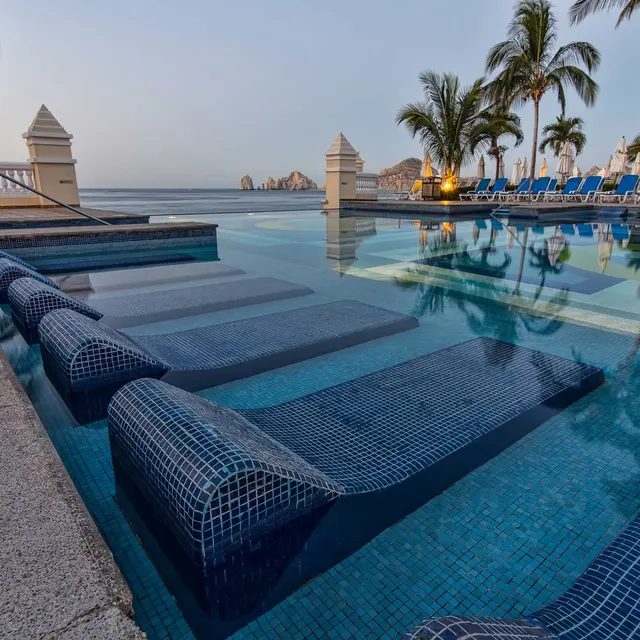 Serene pool with lounge seats, palm trees, and ocean view at dusk, inviting relaxation amidst tranquil surroundings.
