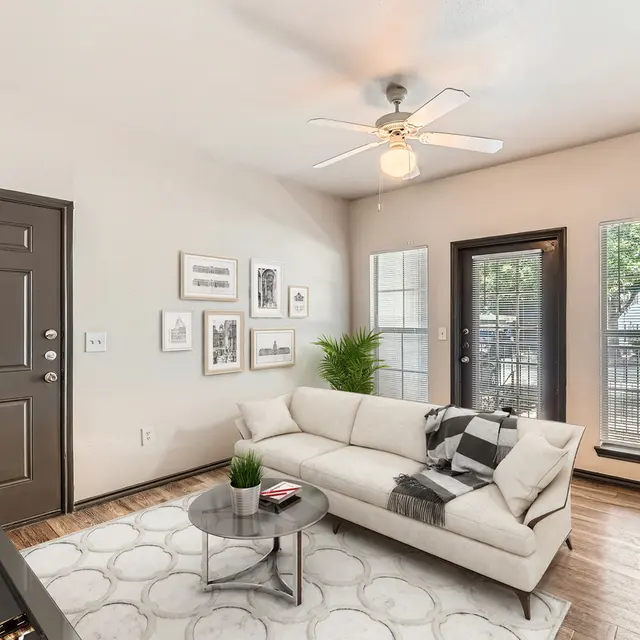 A modern living room featuring a light-colored sofa, a round coffee table, and large windows letting in natural light. The walls are adorned with framed pictures, and there is a plant in the corner.