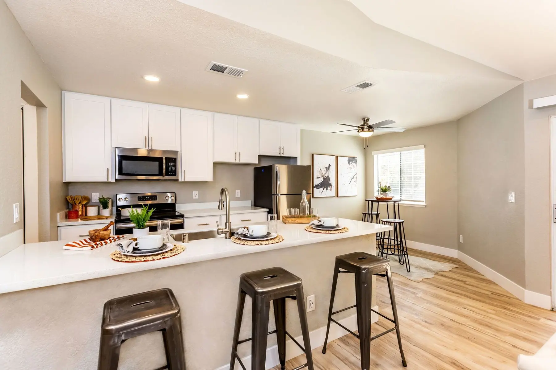 A modern kitchen featuring a white countertop, stainless steel appliances, and a small dining area with bar stools. The kitchen has light gray walls and wooden flooring, creating a bright and inviting atmosphere.