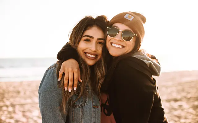 Two smiling women embrace on a beach, wearing casual clothing and enjoying the sunny atmosphere. The ocean is visible behind them.