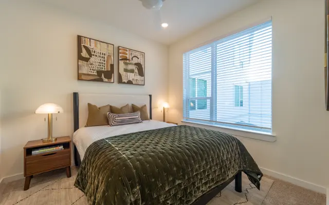 A cozy bedroom featuring a bed with a green quilt, two decorative pillows, and framed artwork on the wall. There is a nightstand with a lamp next to the bed and a large window with blinds allowing natural light to enter.