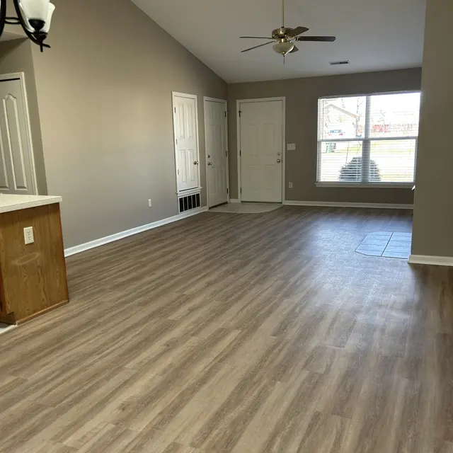 A spacious living room featuring light brown wooden flooring, beige walls, and vaulted ceilings. There are two doors on the left side leading to other rooms, and a large window on the right letting in natural light. A ceiling fan hangs above in the center of the room.