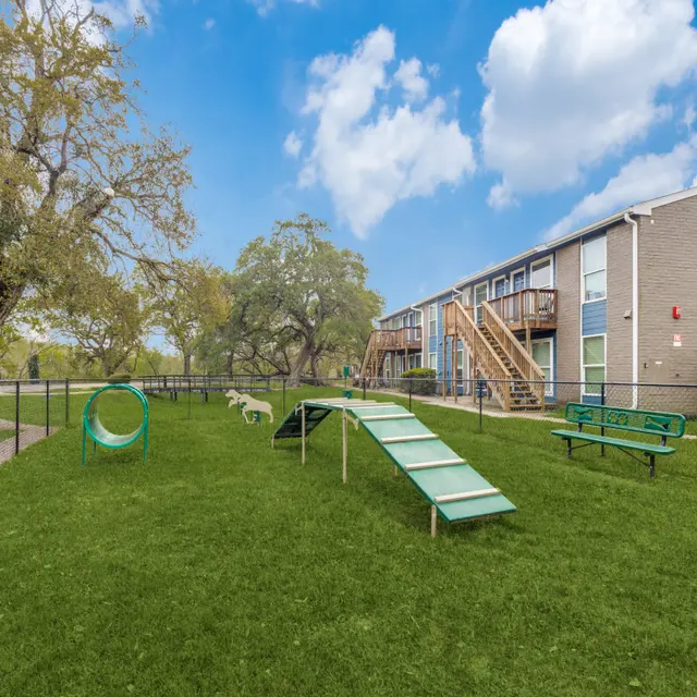 A spacious, fenced dog park featuring green grass, a climbing ramp, a circular play structure, and benches. In the background, an apartment building with a wooden staircase is visible under a blue sky with fluffy clouds.