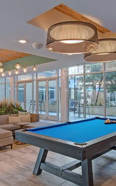 Modern resident clubhouse at 120 Ninth Street Apartments in San Antonio, featuring a pool table, lounge seating, and floor-to-ceiling windows overlooking the courtyard and grill area.