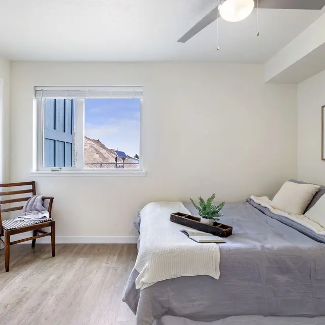 Stylish and Minimalist Bedroom Design A modern bedroom featuring a bed with gray linens, a small wooden chair next to the bed, and a window allowing natural light to enter. A simple piece of artwork is hanging on the wall, and there is a potted plant on the bed.