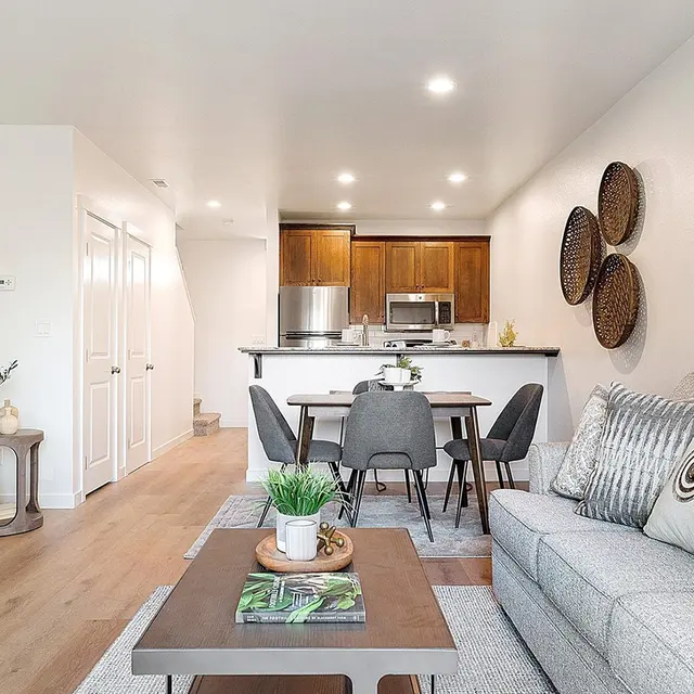 A modern apartment interior featuring a cozy living room with a gray sofa, a coffee table with decorative items, and a dining area. The kitchen is visible in the background with wooden cabinetry and appliances.