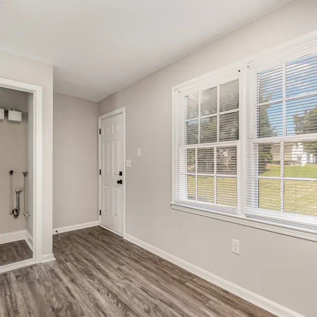 Spacious interior room with light gray walls, hardwood floors, bright windows, and a door leading outside.